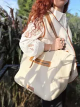 Bolso tipo tote color crema, con detalles en cuero color marrón. Tiene un bolsillo con cierre en la parte superior y un parche de cuero con el logo de la marca en la parte inferior.