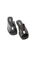 Sandalia plana negra de cuero blando con pala cruzada y aplique metálico decorativo.