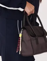Cartera tipo city color beige, de tela deportiva, con doble asa corta, correa regulable desmontable, solapa con broche a presión y charm colgante.