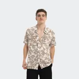 Camisa de manga corta con estampado tropical en tonos beige y blanco, con cuello clásico y cierre frontal con botones.