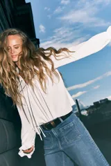 Blusa blanca de manga larga con cuello alto y lazo para atar. Presenta detalles de volados y calados tipo encaje en el canesú y puños con elástico y volado.