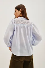 Camisa de algodón color celeste, con escote en V, cierre parcial de botones y detalles de encaje en los hombros. Mangas largas abullonadas con puños elásticos.