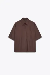 Camisa marrón de popelín con cuello solapa, manga corta con vuelta, bolsillo de plastrón en el pecho, bajo con aberturas laterales, pinzas en la espalda y cierre frontal con botones ocultos.