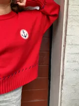 Sweater rojo de punto con cuello redondo y manga larga. Tiene un parche con un ancla bordada en el pecho y detalles de costuras en contraste en puños y ruedo.
