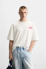 Camiseta blanca de corte relajado, con cuello redondo y mangas cortas. Presenta estampados combinados en contraste en la parte delantera y trasera con la frase "Gourmet Groove LLC".