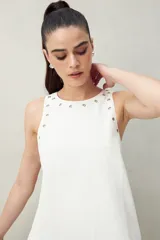 Vestido corto de algodón color verde oliva, de corte recto, con cuello redondo y apliques metálicos en el escote y sisas. Se lleva sobre una camisa negra de manga larga.