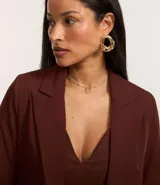 Blazer sastre femenino confeccionado en crepé color marrón, de modelo oversized y comprimento alargado. Sin estampa, posee bolsillos y detalle de pliegues en los puños.