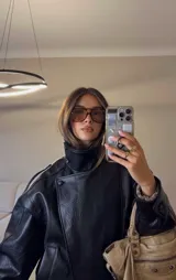 Chaqueta negra de efecto cuero, estilo bomber o motociclista, con cuello alto abotonado y cierre frontal asimétrico. Presenta detalles de costuras verticales y hombreras pequeñas. El bajo es elástico.