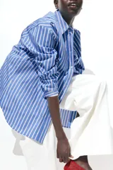 Camisa oversized de popelina de algodón a rayas verticales celestes, blancas y rojas. Tiene cuello clásico, cierre frontal con botones, hombros caídos y mangas largas con puños abotonados. El bajo es redondeado.