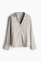 Blusa de manga larga color gris claro o beige pálido, con acabado satinado y cuello clásico que forma un escote en V profundo con cierre de botones frontales.