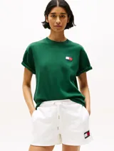 Remera verde de algodón con cuello redondo y corte amplio. Presenta el logo de Tommy Hilfiger en el pecho y un logo oversize en la parte trasera. Tiene mangas cortas con hombros caídos y un parche con logo en la manga.