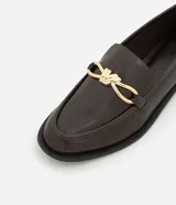 Zapato mocasín de cuero color marrón oscuro con detalle metálico dorado en la parte delantera.