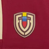Short deportivo Adidas color bordó, con detalles en dorado en las tres franjas laterales y ribete blanco en el borde inferior. Presenta el escudo de la selección de fútbol de Venezuela en la pierna izquierda y el logo de Adidas en la pierna derecha.