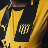 Camiseta de fútbol Puma del Club Atlético Peñarol, modelo Home 2024. Diseño a rayas verticales amarillas y negras con detalles en negro en cuello y mangas. Incluye logo de Puma, escudo del club y sponsor Antel en el frente.