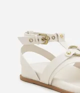 Sandalia plana de cuero sintético color blanco, con tiras superpuestas, detalle de aro metálico y cierre ajustable con hebilla.