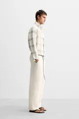Pantalón color crema de lino, corte amplio, con pliegues frontales, bolsillos laterales y traseros de ojal. Cierre frontal con cremallera y gancho metálico oculto.