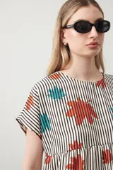 Vestido corto de silueta amplia con corte bajo busto, cuello a la base y mangas cortas. El tejido presenta un estampado de hojas en tonos rojo óxido y turquesa sobre un fondo de rayas verticales blancas y negras.