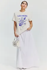 Remera color crudo de algodón con cuello redondo y mangas cortas. Estampado frontal con la frase "LES MERS" y dibujos de conchas y estrellas de mar en color azul.