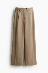 Pantalón sastrero color beige de tiro alto con cintura elástica, cierre y pretina prolongada con botón. Modelo con bolsillos al bies, solapa decorativa con botón adelante y bolsillos de atrás insertados. Piernas amplias con pinzas y pliegues adelante y atrás.