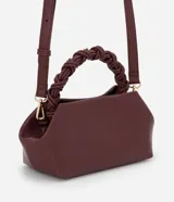 Cartera pequeña color borgoña, con asa corta trenzada y correa larga ajustable y desmontable. Cierre superior con broche imantado. Interior forrado con bolsillo.