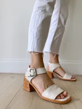 Sandalias de cuero color crudo, con tira ancha sobre los dedos, pulsera al tobillo con hebilla metálica y taco de madera de 6 cm de altura.