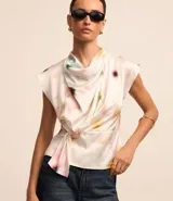 Blusa sin mangas de tela satinada color blanco, con cuello drapeado, lazo lateral y estampado de manchas en tonos rosados, amarillos y verdes.