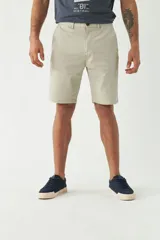 Bermuda color beige de gabardina, corte chino, con bolsillos laterales y traseros. Tiene pasacinto, cierre con botón y bragueta, y logo metálico pequeño en el frente.