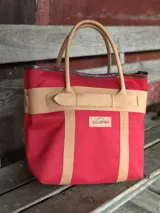 Bolso de playa de lona color verde claro con detalles en cuero color beige. Tiene doble asa de hombro y cierre superior con tira de cuero.