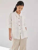 Camisa negra de lino con corte femenino, cuello clásico, cierre frontal con botones y mangas 3/4 con puños enrollados. Presenta detalles de frunces verticales en el frente y bolsillos de parche a la altura de la cadera.