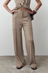 Pantalón de sarga color beige, de tiro medio con cintura elástica en la espalda, bolsillos laterales de plastrón con botón metálico y pierna ancha.