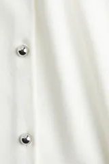 Chaleco corto de punto color blanco, con cuello redondo y cierre frontal con botones metálicos.