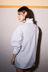 Camisa celeste a rayas verticales azules y marrones, con cuello blanco, cierre frontal con botones dorados y volantes verticales.