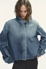 Chaqueta de denim celeste con cuello alto, manga larga con trabilla y bolsillos laterales. Presenta detalle de pliegues en la espalda y cierre frontal con botones ocultos.