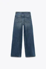 Pantalón de jean celeste de tiro alto, corte wide leg y largo hasta el tobillo. Presenta un diseño asimétrico en la cintura con cierre frontal cruzado con cremallera y botones metálicos.