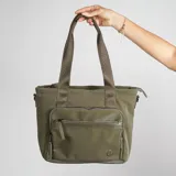 Bolso tipo tote bag color verde militar, con doble asa de hombro y cierre superior. Tiene un bolsillo frontal con cierre y logo circular de la marca en la esquina inferior.