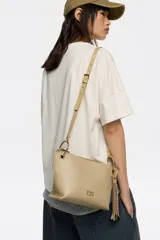 Bolso bandolera de piel vacuna negra, con forma trapezoidal y estructura rígida. Posee asa regulable, cierre de cremallera, bolsillo interior y charm decorativo con borlas en tonos tierra y dorado.