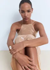 Vestido midi color beige, con diseño recto y escote halter con cierre de lazo en la espalda. El tejido principal es de lentejuelas.