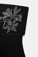 Body palabra de honor negro confeccionado en tejido elástico, con bordados florales y aplicación de brillos en la parte superior. Cierre en bajo con botones a presión.