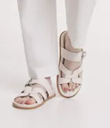 Sandalias planas de cuero sintético color blanco, con puntera redonda y dos tiras, una con hebilla metálica y otra en forma de H.