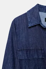 Vestido midi camisero de denim azul con cuello solapa y escote pico. Manga larga, bolsillos de parche con solapa en el delantero y laterales ocultos en costura. Incluye cinturón de la misma tela y cierre frontal con botones ocultos.