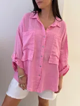 Camisa oversize rosada de lino, con cuello clásico, cierre frontal con botones, dos bolsillos de parche en el pecho y mangas largas con puños abotonados.