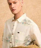Camisa de manga corta con estampado de palmeras y motivos playeros en tonos celeste y verde sobre fondo blanco.