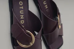 Sandalia plana de cuero, color marrón oscuro, con tira ancha cruzada sobre el empeine y hebilla metálica dorada.