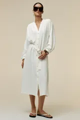 Vestido midi blanco de crepe con cuello redondo y escote pico, manga larga con puños y cinturón de la misma tela con hebilla metálica. Cierre frontal con botones metálicos.