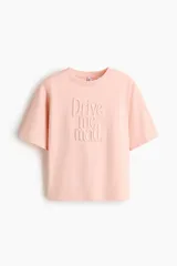 Camiseta de punto suave de algodón color rosado pálido, con corte holgado y mangas cortas. Presenta un motivo con texto en relieve/bordado en el frente que dice "Drive me mad".