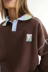 Buzo marrón con cuello polo celeste con rayas amarillas, mangas largas con costuras a contratono y parche bordado en el pecho.