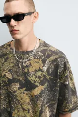 Sudadera relaxed fit con estampado de hojas en tonos verde, marrón y negro. Tiene cuello redondo, manga corta y bajo irregular con aberturas laterales.