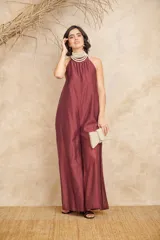 Mono de seda fluida color borgoña, con corte amplio, espalda descubierta y cuello halter con cierre de lazo. Incluye un collar de perlas de tres vueltas y un clutch tejido color crema.