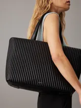 Bolso tote negro Calvin Klein, confeccionado en piel sintética acolchada con diseño de rayas verticales. Tiene doble asa de hombro, cierre superior con cremallera, herrajes plateados y logo metálico en el frente.