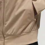 Campera Adidas Originals Adicolor Classics Firebird color beige con tres rayas blancas en las mangas, logo bordado en el pecho, cuello alto, cierre frontal y bolsillos con cremallera.